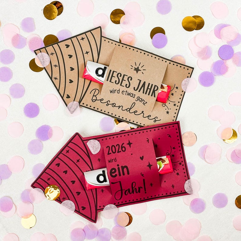 Puede incluir: Dos tarjeteros de regalo con caramelos, rodeados de confeti. Uno es marr&oacute;n con un dise&ntilde;o de coraz&oacute;n y estrella, el otro es rojo con un dise&ntilde;o de coraz&oacute;n. La tarjeta marr&oacute;n dice "DIESES JAHR wird etwas ganz Besonderes". La tarjeta roja dice "2026+ wird dein Jahr!"