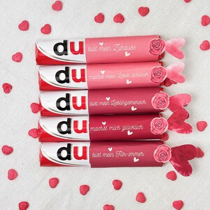 Pu&ograve; includere: Quattro barrette di cioccolato con confezione rossa e bianca, ciascuna avvolta in una diversa tonalit&agrave; di carta rosa. Le confezioni hanno testo in tedesco e sono decorate con rose di carta e coriandoli a forma di cuore.