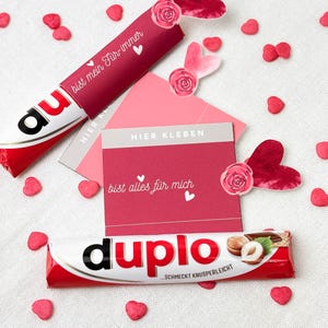 Pu&ograve; includere: Una composizione a tema San Valentino con una barretta di cioccolato rossa e bianca, un involucro di caramelle rosso con il testo "bist mein F&uuml;r-immer" e una carta rosa con il testo "bist alles f&uuml;r mich". Cuori decorativi e rose.