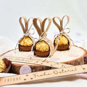 May include: Three Easter-themed treats on a wooden slice. Each treat is a gold-wrapped chocolate in a brown paper cup, adorned with brown paper bunny ears and tied with twine. A banner with the words "Frohe Ostern!" is in the foreground.