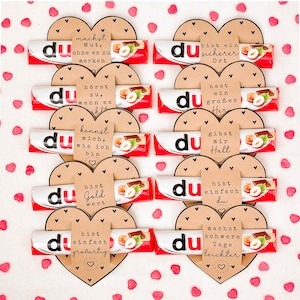Puede incluir: Arreglo de barras de chocolate con la palabra "du" en el envoltorio, dispuestas en forma de corazón con frases alemanas como "Haces coraje" y "Eres simplemente genial". Envoltorios rojos y blancos.
