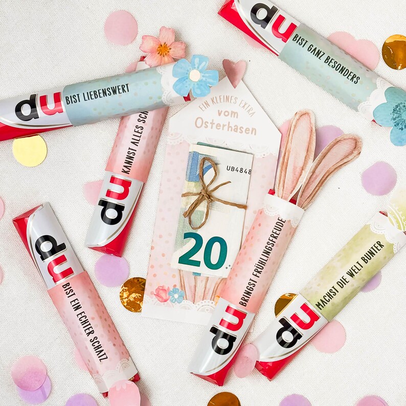 May include: A collection of colorful candy bars with German text, scattered with confetti and decorative elements. Some bars have text like "Bist Liebenswert" and "Bist ein echter Schatz." A card with money and the number 20 is also present.