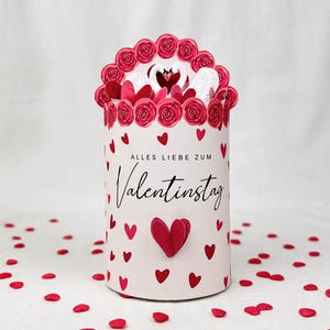 Pu&ograve; includere: Una scatola regalo cilindrica di San Valentino con una parte superiore floreale e dettagli a forma di cuore. La scatola &egrave; decorata con cuori e rose rosse, e il testo "ALLES LIEBE ZUM Valentinstag" &egrave; scritto sul davanti.
