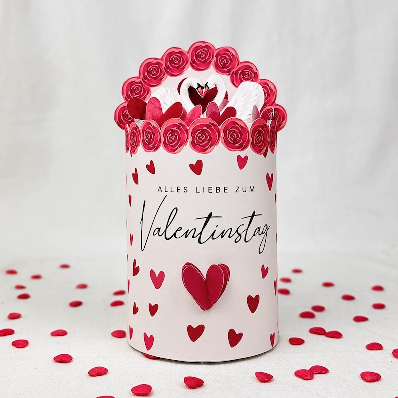Pu&ograve; includere: Una scatola regalo cilindrica di San Valentino con una parte superiore floreale e dettagli a forma di cuore. La scatola &egrave; decorata con cuori e rose rosse, e il testo "ALLES LIEBE ZUM Valentinstag" &egrave; scritto sul davanti.