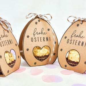 Puede incluir: Tres cajas de regalo de papel marr&oacute;n con forma de huevo de Pascua con una ventana en forma de coraz&oacute;n que revela un caramelo de chocolate. Cada caja tiene el texto alem&aacute;n "Frohe OSTERN!" y est&aacute; decorada con peque&ntilde;as estrellas y un lazo de cuerda.