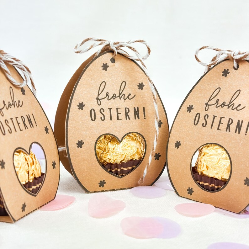 Puede incluir: Tres cajas de regalo de papel marr&oacute;n con forma de huevo de Pascua con una ventana en forma de coraz&oacute;n que revela un caramelo de chocolate. Cada caja tiene el texto alem&aacute;n "Frohe OSTERN!" y est&aacute; decorada con peque&ntilde;as estrellas y un lazo de cuerda.
