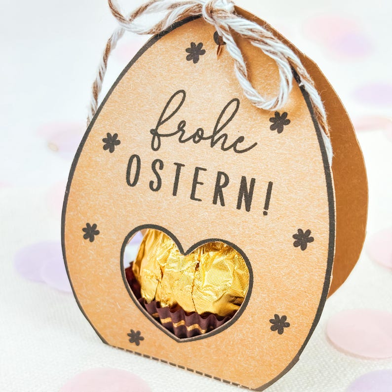 Puede incluir: Una caja de regalo marr&oacute;n con forma de huevo de Pascua con una ventana en forma de coraz&oacute;n que revela un chocolate dorado. La caja tiene las palabras "Frohe OSTERN!" y dise&ntilde;os de estrellas. Un lazo de cuerda est&aacute; en la parte superior.