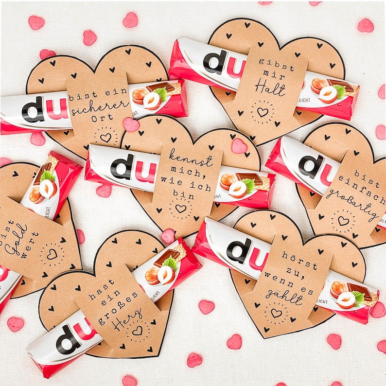 May include: A collection of heart-shaped cards with various German phrases and small heart illustrations. Each card holds a chocolate bar with the word "du" on it. Pink heart confetti is scattered around the cards.