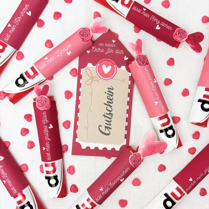 Pu&ograve; includere: Un assortimento di barrette di caramelle avvolte in carta rossa e rosa, decorate con cuori e rose. Le barrette hanno testo in tedesco e sono disposte attorno a un'etichetta regalo con la scritta "Gutschein".