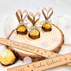 May include: Three Easter-themed chocolate candies wrapped in gold foil and brown paper, shaped like bunnies, sit on a wooden slice. The candies are tied with twine and surrounded by confetti and a banner that says "Frohe Ostern!"