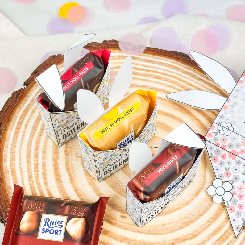 Puede incluir: Tres barras de chocolate Ritter Sport en cajas decorativas con detalles de orejas de conejo y una barra sin envolver. Las cajas est&aacute;n decoradas con motivos florales y el texto "Frohe Ostern". Los chocolates est&aacute;n sobre una superficie de madera.