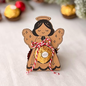May include: A die-cut brown cardboard angel ornament featuring black line art of a smiling face and wings, holding a gold-foil wrapped Ferrero Rocher chocolate in the center, secured with a red and white twisted twine bow tied at the waist.
