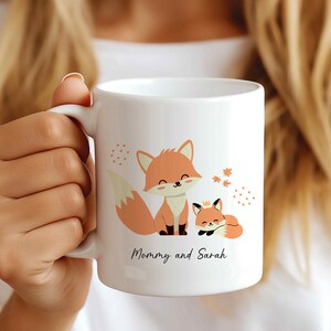 May include: White ceramic mug with a design of two foxes. The larger fox is standing and the smaller fox is lying down. The text "Mommy and Sarah" is printed on the mug.