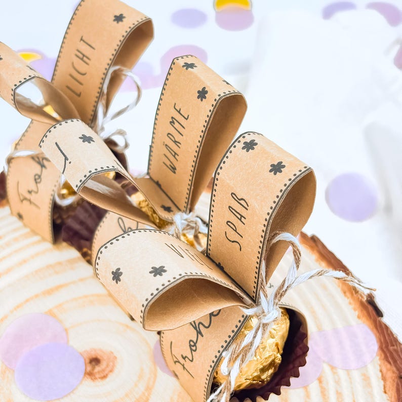 May include: Close-up of wrapped candies with brown paper bows. Each bow has a word printed on it, including "Licht", "W&auml;rme", and "Spa&szlig;". The candies are tied with twine and decorated with small star-shaped designs. The background includes confetti.