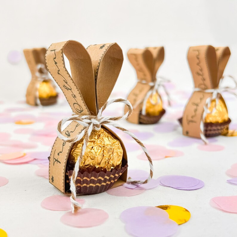 May include: Several Easter-themed candy packages. Each package features a gold-wrapped chocolate candy in a brown paper basket, wrapped in brown paper with bunny ear accents. The packages are tied with twine and sit on a surface scattered with confetti.