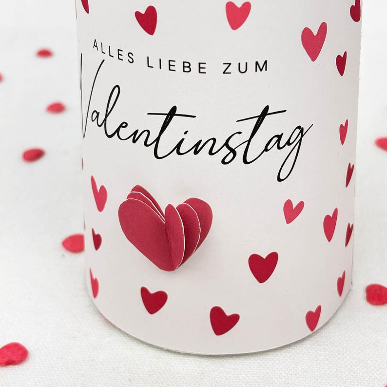 Pu&ograve; includere: Un cilindro bianco decorato con cuori rossi e la scritta "Alles Liebe zum Valentinstag" in corsivo nero. Un cuore di carta 3D &egrave; attaccato al cilindro. Coriandoli a forma di cuore rosso sparsi.