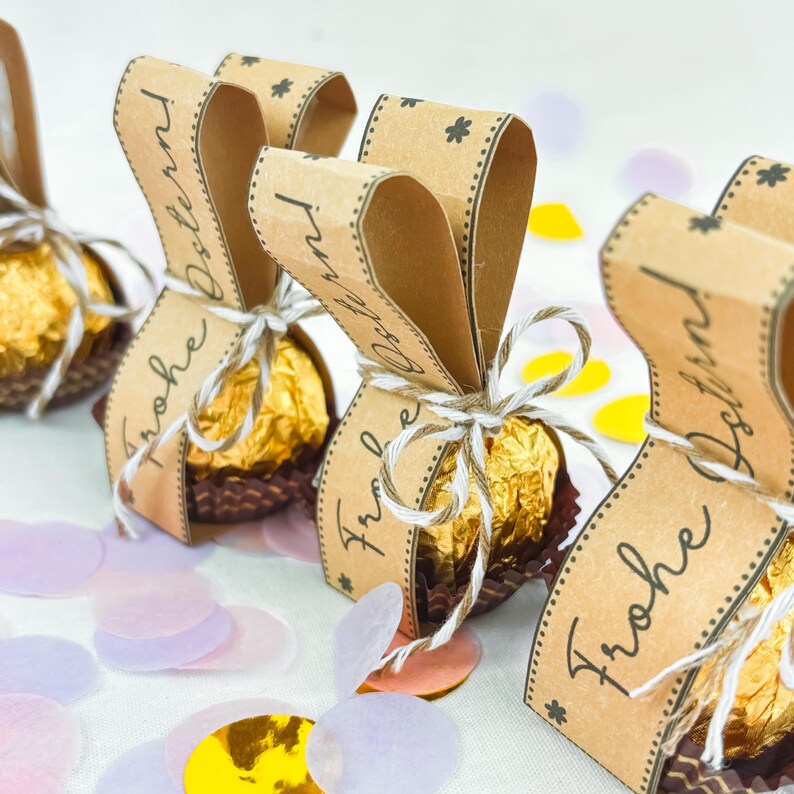 May include: Close-up of Easter-themed chocolate candies wrapped in gold foil and presented in decorative brown paper packaging. Each package is tied with twine and adorned with a brown paper tag that reads "Frohe Ostern". Confetti in pastel colors surrounds the candies.