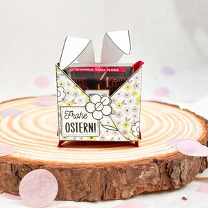 Puede incluir: Una caja de regalo decorativa con un estampado floral y el texto "Frohe Ostern!" se muestra sobre una superficie de madera. La caja contiene barras de chocolate. La caja est&aacute; decorada con recortes de papel blanco.
