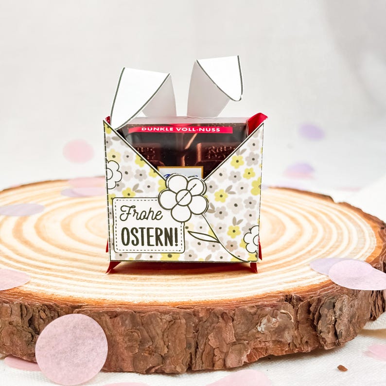 Puede incluir: Una caja de regalo decorativa con un estampado floral y el texto "Frohe Ostern!" se muestra sobre una superficie de madera. La caja contiene barras de chocolate. La caja est&aacute; decorada con recortes de papel blanco.