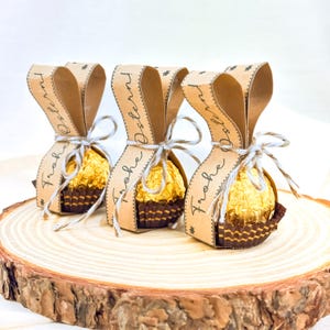 May include: Three Easter-themed gift packages on a wooden slice. Each package features a gold-wrapped chocolate candy in a brown paper cup, tied with twine, and a brown paper ribbon printed with "Frohe Ostern".