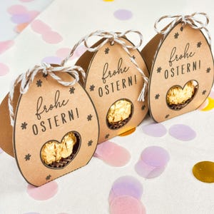 Puede incluir: Tres cajas de regalo marrones con forma de huevo de Pascua con el texto "Frohe OSTERN!" y estrellas. Cada caja tiene una ventana en forma de coraz&oacute;n que revela un chocolate envuelto en oro. Las cajas est&aacute;n atadas con hilo y sobre una superficie cubierta de confeti.