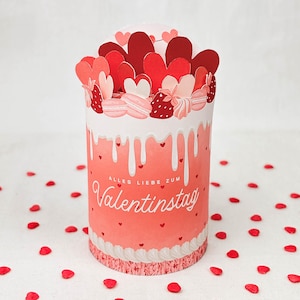 May include: A cylindrical Valentine's Day gift box with a red and white design. The box is decorated with red hearts, macarons, and the text "Alles Liebe zum Valentinstag". The box is surrounded by red heart-shaped confetti.
