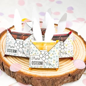 Puede incluir: Tres cajas de caramelos con tem&aacute;tica de Pascua con orejas de conejo blancas, colocadas sobre una rodaja de madera. Las cajas est&aacute;n decoradas con motivos florales y el texto "Frohe OSTERN!". Cada caja contiene una barra de chocolate.
