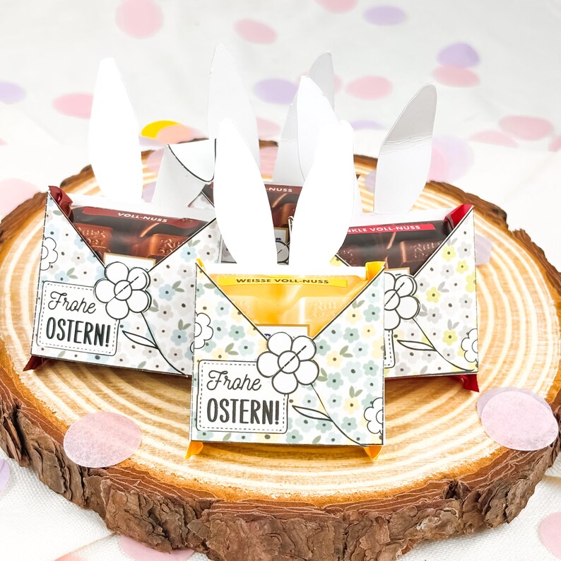 Puede incluir: Tres cajas de caramelos con tem&aacute;tica de Pascua con orejas de conejo blancas, colocadas sobre una rodaja de madera. Las cajas est&aacute;n decoradas con motivos florales y el texto "Frohe OSTERN!". Cada caja contiene una barra de chocolate.
