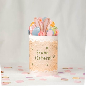 May include: A decorative Easter-themed container with a woven basket design. The container is adorned with paper cutouts of Easter eggs, flowers, and bunny ears. The text "Frohe Ostern!" is printed on the front.