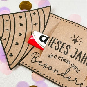 Puede incluir: Dos tarjeteros de papel marr&oacute;n con l&iacute;neas decorativas negras y texto. Un soporte tiene una barra de caramelo asomando. El texto de la tarjeta dice "DIESES JAHR wird etwas ganz Besondere". Confeti dorado, rosa y morado esparcido.