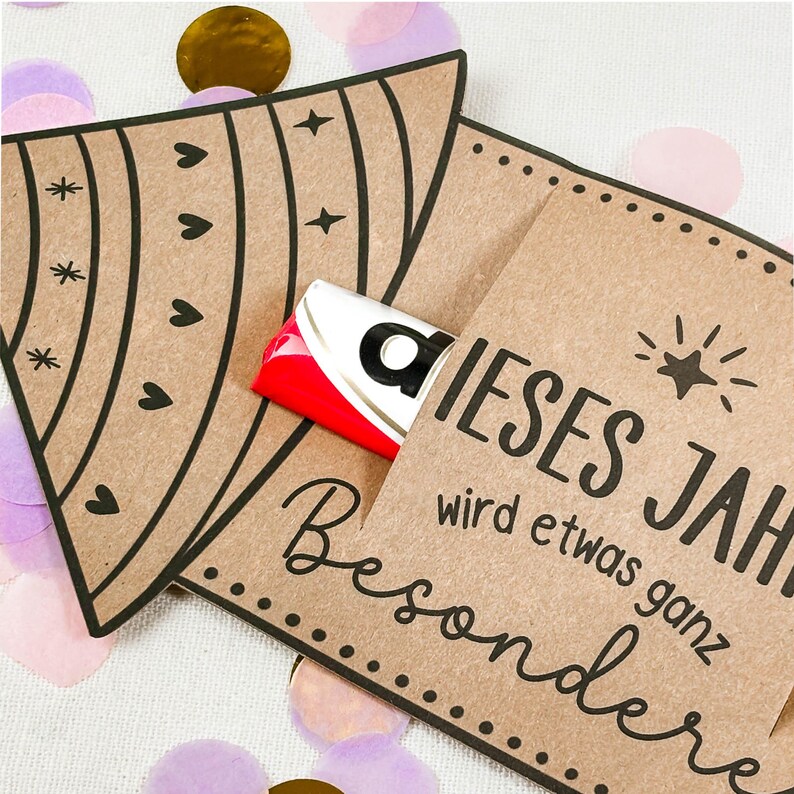 Puede incluir: Dos tarjeteros de papel marr&oacute;n con l&iacute;neas decorativas negras y texto. Un soporte tiene una barra de caramelo asomando. El texto de la tarjeta dice "DIESES JAHR wird etwas ganz Besondere". Confeti dorado, rosa y morado esparcido.