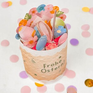 May include: A decorative Easter basket with a woven texture, filled with colorful paper cutouts of eggs, flowers, and bunny ears. The basket has a white lace trim and the words "Frohe Ostern!" are printed on the side. Confetti in various colors surrounds the basket.