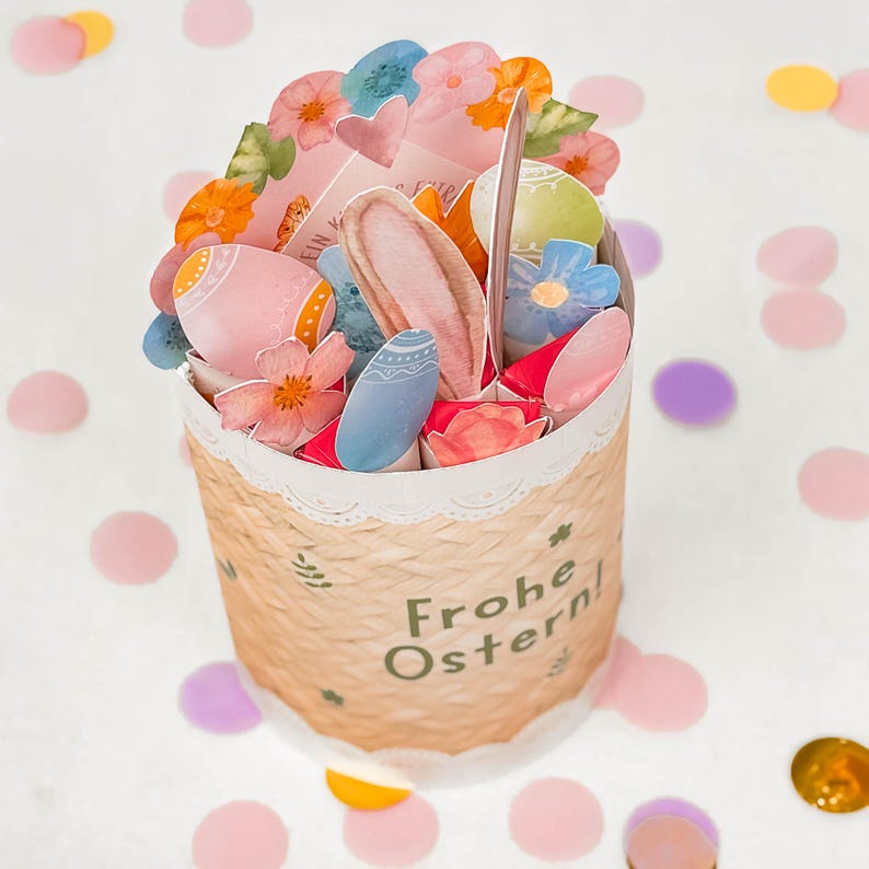 May include: A decorative Easter basket with a woven texture, filled with colorful paper cutouts of eggs, flowers, and bunny ears. The basket has a white lace trim and the words "Frohe Ostern!" are printed on the side. Confetti in various colors surrounds the basket.