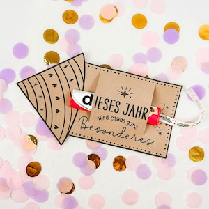 Puede incluir: Una tarjeta de regalo de papel kraft con el texto alem&aacute;n "DIESES JAHR wird etwas ganz Besonderes" y una barra de chocolate Kinder. La tarjeta est&aacute; decorada con corazones y estrellas negras, y una cinta. Confeti dorado, rosa y morado rodea la tarjeta.
