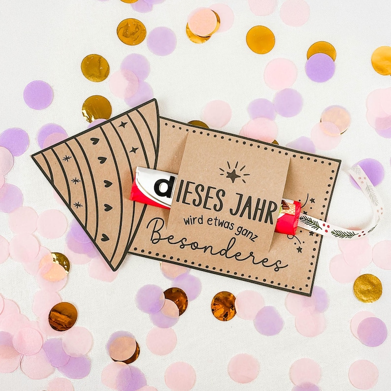 Puede incluir: Una tarjeta de regalo de papel kraft con el texto alem&aacute;n "DIESES JAHR wird etwas ganz Besonderes" y una barra de chocolate Kinder. La tarjeta est&aacute; decorada con corazones y estrellas negras, y una cinta. Confeti dorado, rosa y morado rodea la tarjeta.