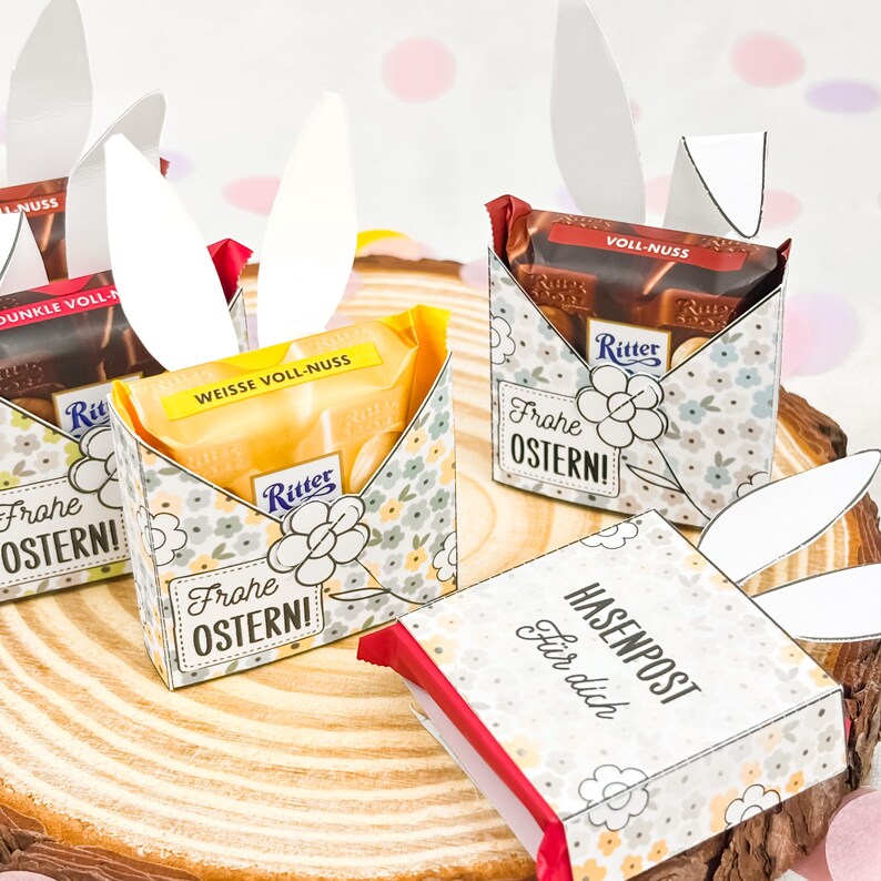 Puede incluir: Cajas de regalo de Pascua con barras de chocolate, dise&ntilde;os florales y orejas de conejo de papel. Las cajas tienen el texto "Frohe OSTERN!" y "HASENPOST F&uuml;r dich". Chocolates Ritter.