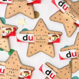 May include: Several star-shaped kraft paper holders each containing a red and white chocolate bar. The paper stars feature black printed star motifs and German text including 'du bist ein Glücksfall', 'du bist ein heller Stern', and 'du versüßt Weihnachten'.