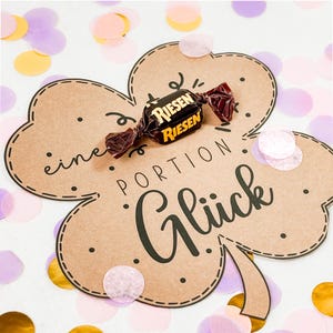 May include: A brown paper clover with the words "eine Portion Gl&uuml;ck" and a wrapped Riesen candy. The clover is decorated with small black dots and surrounded by confetti in shades of pink, yellow, and purple.