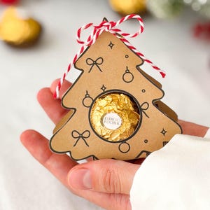 May include: A brown cardboard Christmas tree-shaped gift box featuring black line drawings of bows and ornaments. A single gold-wrapped Ferrero Rocher chocolate is visible through a circular cutout in the center. The box is tied with red and white twine.