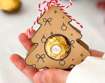May include: A brown cardboard Christmas tree-shaped gift box featuring black line drawings of bows and ornaments. A single gold-wrapped Ferrero Rocher chocolate is visible through a circular cutout in the center. The box is tied with red and white twine.