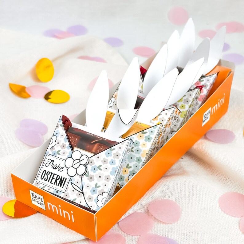 Puede incluir: Una caja naranja y blanca de chocolates Ritter Sport Mini, decorada para Semana Santa. La caja presenta orejas de conejo blancas y envoltorios de chocolate con motivos florales. La caja tiene el texto "Frohe Ostern!" y "mini".