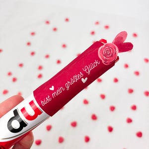 Pu&ograve; includere: Una barretta di cioccolato rossa e bianca con la parola "Nutella" visibile. La barretta &egrave; avvolta in carta rossa con la frase tedesca "bist mein gr&ouml;&szlig;tes Gl&uuml;ck" e piccoli cuori bianchi. &Egrave; attaccata una rosa di carta e un ornamento a forma di cuore.