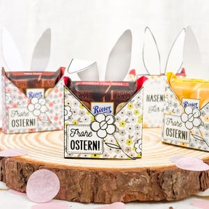 Puede incluir: Cajas de regalo de Pascua con forma de conejos, cada una con una barra de chocolate Ritter. Las cajas est&aacute;n decoradas con motivos florales y llevan la inscripci&oacute;n "Frohe Ostern!". Se exhiben sobre una superficie de madera.