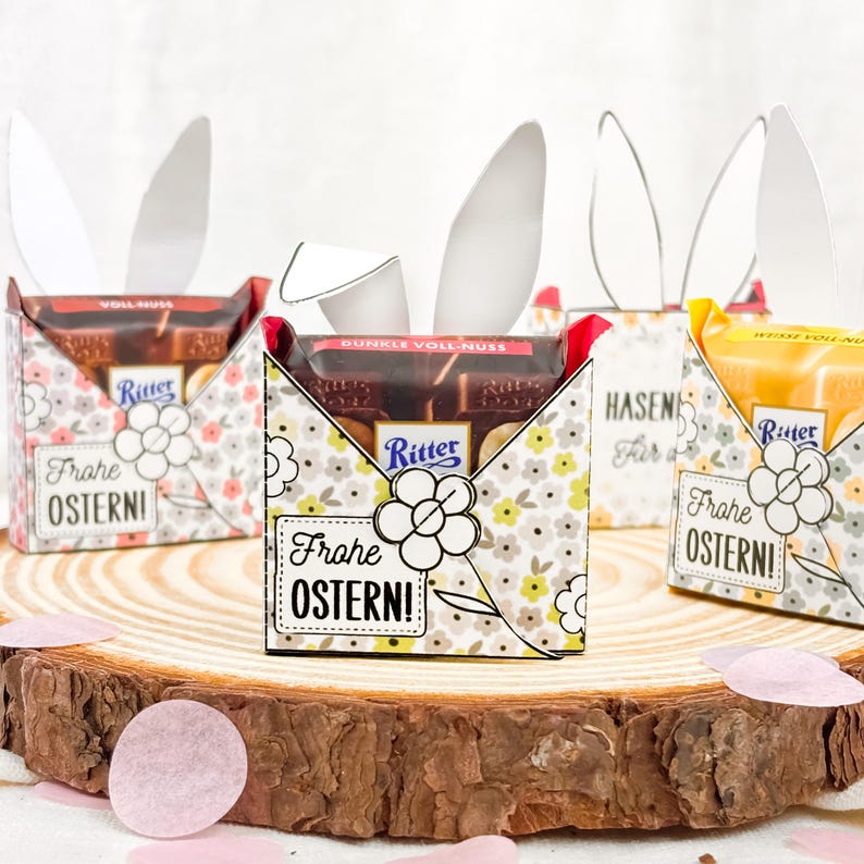 Puede incluir: Cajas de regalo de Pascua con forma de conejos, cada una con una barra de chocolate Ritter. Las cajas est&aacute;n decoradas con motivos florales y llevan la inscripci&oacute;n "Frohe Ostern!". Se exhiben sobre una superficie de madera.