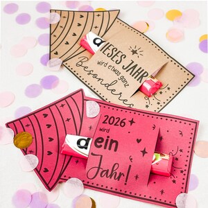 Puede incluir: Dos tarjetas de felicitaci&oacute;n con barras de caramelo. Una tarjeta es marr&oacute;n con el texto "DIESES JAHR wird etwas ganz Besonderes". La otra tarjeta es rosa con el texto "2026 wird ein Jahr!". Confeti de varios colores rodea las tarjetas.