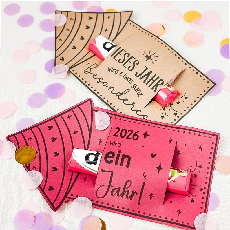 Puede incluir: Dos tarjetas de felicitaci&oacute;n con barras de caramelo. Una tarjeta es marr&oacute;n con el texto "DIESES JAHR wird etwas ganz Besonderes". La otra tarjeta es rosa con el texto "2026 wird ein Jahr!". Confeti de varios colores rodea las tarjetas.