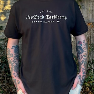 May include: Black t-shirt with the text "LivDead Taxidermy" in a white, elegant font. The text includes "EST 2024" above and "Grand Rapids, MI" below. The shirt is a classic crew neck style.