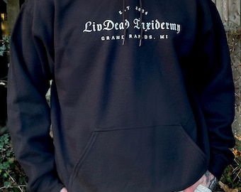 May include: Black hoodie with a front pocket and drawstring hood. The white text on the front reads "LivDead Taxidermy" with "E.T. 2034" above and "Grand Rapids, MI" below. The person is wearing khaki pants.
