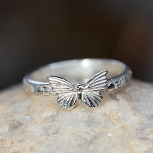 May include: A silver ring with a butterfly design. The butterfly is the focal point, with detailed wing patterns. The ring band has a textured pattern. The ring is displayed on a light-colored stone.