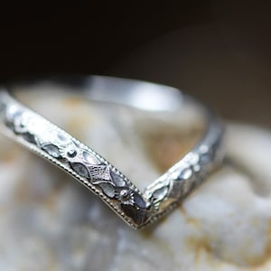 May include: A silver V-shaped band with an intricate floral design. The band is polished and has a smooth finish.